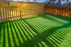 shadows of  a wooden picket fence in a front yard, front garden with artifical grass as a lawn and a red brick perimiter wall.
